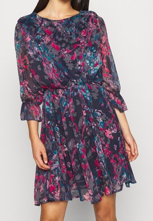 Frau in einem knielangen, dunkelblumenmuster Kleid mit langen, transparenten Ärmeln und einer taillierten Taille.