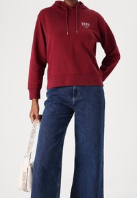 Sudadera burdeos con cordón, que presenta un pequeño logo blanco. Combinada con vaqueros de denim anchos de color azul y un bolso crossbody crema con una correa con logo.