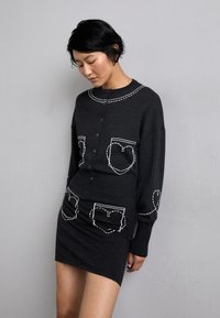 Set de cardigan și fustă din tricot negru, cu detalii albe la buzunare, guler rotund și închidere cu nasturi, având o siluetă ajustată.