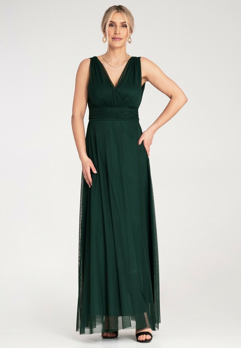 Długa, szmaragdowo-zielona sukienka z lejącego materiału; ma plisowany gorset, dekolt w kształcie litery V i przezroczystą nakładkę. Czarne szpilki dopełniają stylizację.
