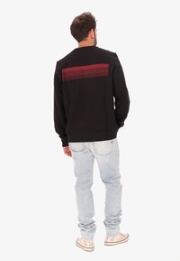 Un uomo sta in piedi di spalle, indossando una felpa nera con una striscia orizzontale rossa, jeans azzurri chiari e sneakers alte beige su uno sfondo bianco.