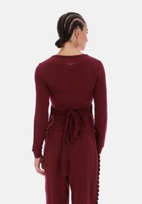 Bordeauxrode top met lange mouwen, voorzien van een strik in de taille, een gladde textuur en een ronde halslijn. Logo gedrukt op de achterkant, met een aansluitend ontwerp.