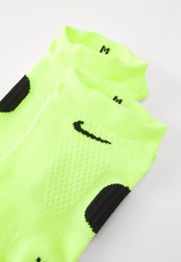Ein Paar neon-gelbe Knöchelstrümpfe mit schwarzen Nike-Swoosh-Logos und schwarzen Fersenakzenten auf weißer Oberfläche.