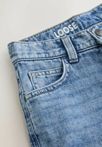 Vapaat siniset farkut, joissa on yksi etutasku, ommellut saumat ja näkyvä metallinappula vyötäröllä.