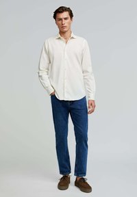 Weißes Hemd mit langen Ärmeln, kombiniert mit blauen Jeans und braunen Schnürschuhen, vor einem hellgrauen Hintergrund stehend.