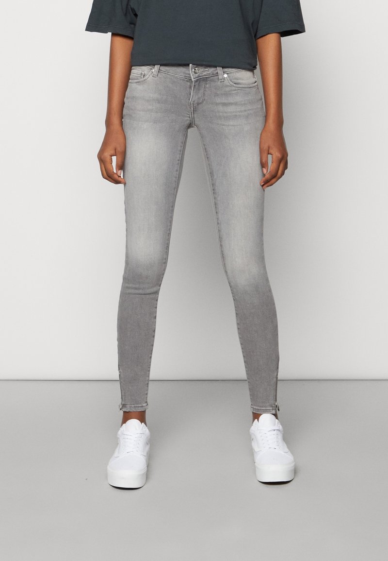 Personne portant un jean skinny gris délavé et des baskets blanches, debout sur un sol gris uni contre un mur blanc.