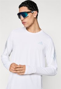 Λευκό πουκάμισο μακριάς μανίκας με μπλε ρίγες στα μανίκια, συνδυασμένο με μεγάλα καθρεπτιστά γυαλιά ηλίου με μαύρο πλαίσιο και διακοσμητικές λεπτομέρειες.