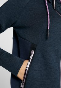 Sudadera azul marino con un tejido texturizado, que presenta un cierre frontal con cremallera, acentos oscuros en contraste y una capucha con cordones rosas.