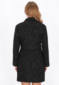 Cappotto nero con texture e motivo intrecciato, maniche lunghe e colletto, con sottili accenti metallici distribuiti. Lunghezza fino a metà coscia.