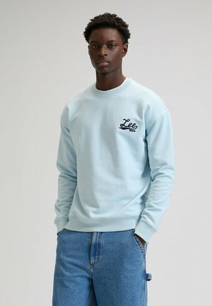 Lichtblauwe sweatshirt met geribde ronde hals en manchetten. Heeft een zwart geborduurd logo op de linkerborst. Gecombineerd met blauwe spijkerbroek.