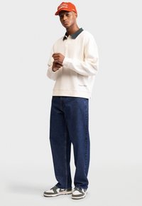 White sweatshirt with dark green collar, paired with dark blue baggy jeans and gray sneakers. Red cap with white text. Detail: embroidered logo.