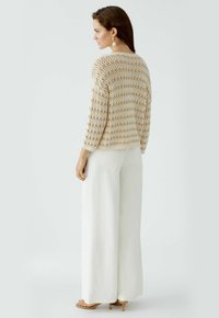 Strickpullover mit offener Webart in Creme- und Brauntönen, kombiniert mit hoch taillierten weißen weiten Hosen. Minimale Hardware und keine sichtbaren Muster.