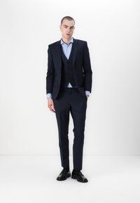 Donker marineblauw pak met een getailleerde blazer en bijpassende broek, aangevuld met een lichtblauwe overhemd. Zwarte schoenen maken het geheel af.