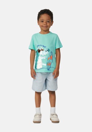 T-shirt en coton vert menthe avec un design en sequins représentant un dinosaure surfeur, associé à un short en denim clair et des baskets blanches.