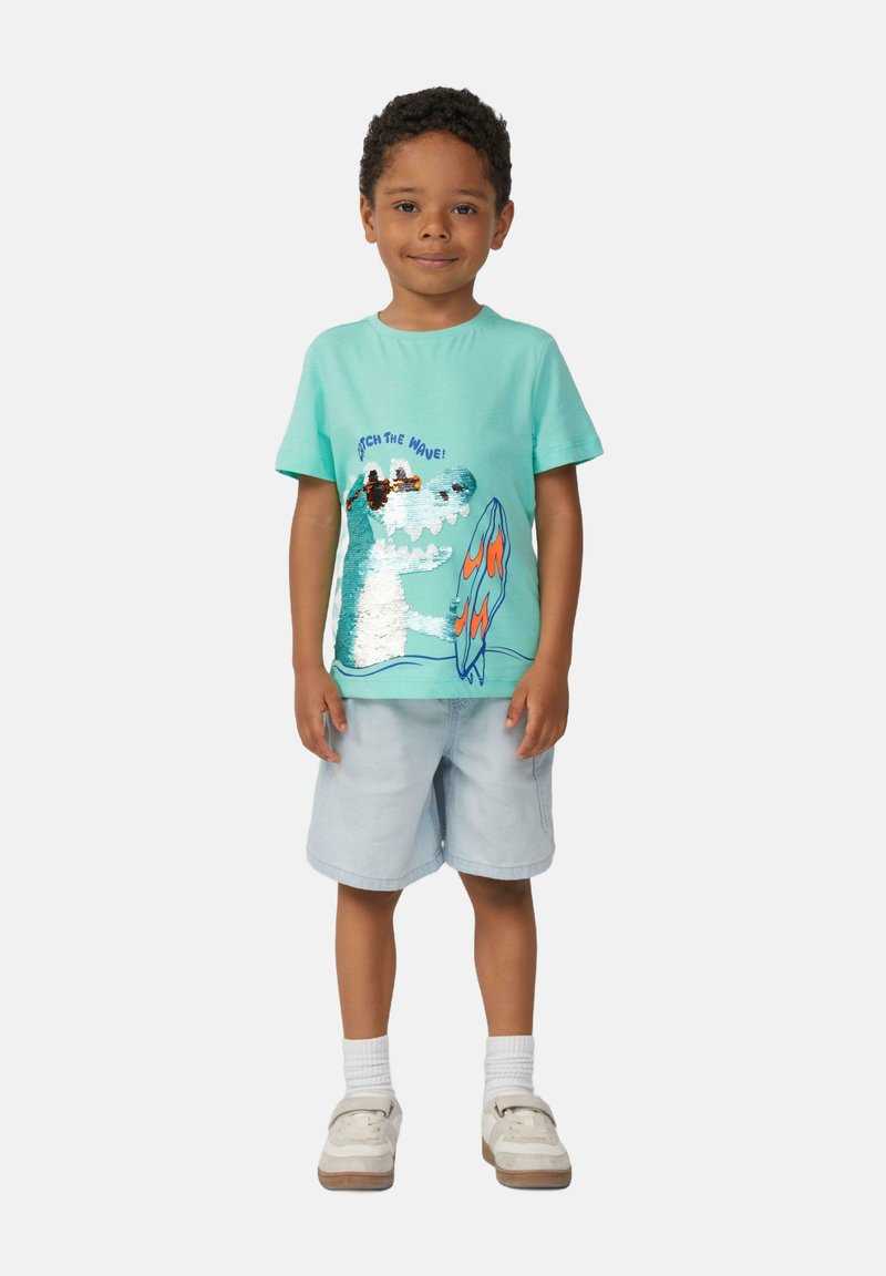 Miętowo-zielona bawełniana koszulka z przyszywanym wzorem przedstawiającym dinozaura surfującego, w zestawie z jasnoniebieskimi szortami dżinsowymi i białymi sneakersami.
