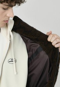 Veste en polaire marron avec une doublure sombre, dotée d'une fermeture éclair et d'une capuche crème en dessous. La capuche comporte un texte imprimé.