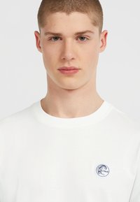 Hvid bomulds t-shirt med rund halsudskæring og et cirkulært blåt logo-patch på venstre bryst. Glat tekstur, afslappet pasform.