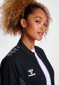 Schwarze Zip-Jacke mit grauen Akzenten und einem strukturierten Streifendesign auf den Schultern. Hat ein Logo in weiß auf der linken Brustseite.
