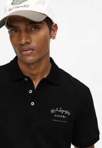 Polo noir en tissu texturé, avec une patte de boutonnage à trois boutons et un logo brodé en argent. Porté avec une casquette beige et blanche.