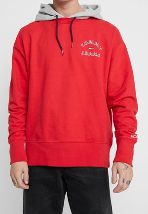 Mann trägt roten Tommy Jeans Sweatshirt mit grauer Kapuze und schwarzen Kordeln, kombiniert mit schwarzen Hosen, sichtbare Handtattoos.