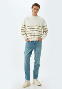 Sudadera de cuello redondo en crema con rayas caqui, combinada con jeans azul claro y zapatillas crema, creando un look casual.