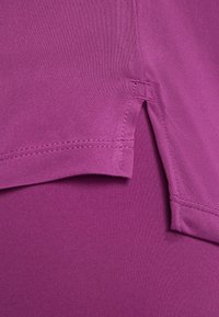 Lila sporttop med slät tyg, rak skärning och sidoslitsar för rörlighet. Sömmen är synlig längs fållen och sömmarna.