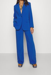 Woman wearing a bright blue oversized suit with wide-leg trousers and a chunky chain necklace, paired with glittery platform shoes.