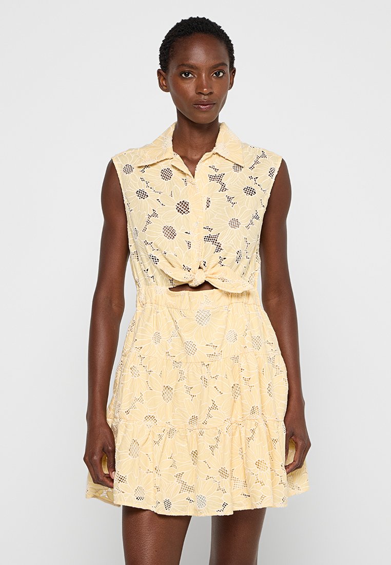 Vestido amarillo sin mangas con diseño de encaje floral, frente anudado y falda en capas. Presenta un collar y sutiles acentos con patrón.