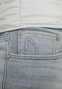 Ljust grå denimficka med metallnitar och en broderad bågformad design, delvis täckt av en vit vadderad jacka.