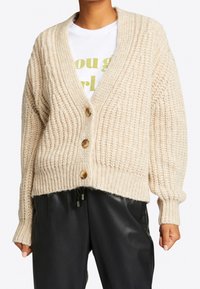 Beżowy sweter dzianinowy z głębokim dekoltem w kształcie V, z zapięciem na guziki, fakturowanym wzorem i ściągaczami przy mankietach. Noszony na białej koszuli z zielonym napisem.