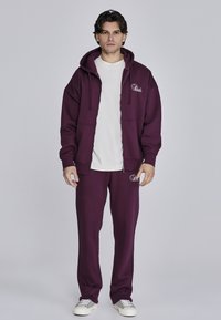 Maroon zip-up dukserica s džepovima, bijela majica ispod, odgovarajuće maroon trenirke i bijele tenisice, s detaljima logotipa na odjeći.