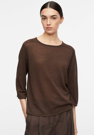 Kobieta z ciemnymi włosami związanymi do tyłu, ubrana w luźną brązową bluzkę z długim rękawem i brązowe spodnie, z rękami w kieszeniach na białym tle.