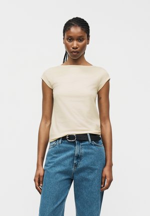 Γυναίκα με πλεγμένα μαλλιά που φοράει μπεζ τοπ με κοντά μανίκια, στερεωμένο μέσα σε ψηλόμεσο μπλε τζιν με μαύρη ζώνη, μπροστά σε απλό λευκό φόντο.