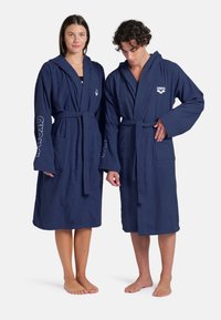 Accappatoi in cotone blu navy con cappuccio e cintura, dotati di due tasche frontali e dettagli con logo sulle maniche. Tessuto leggero e testurizzato.