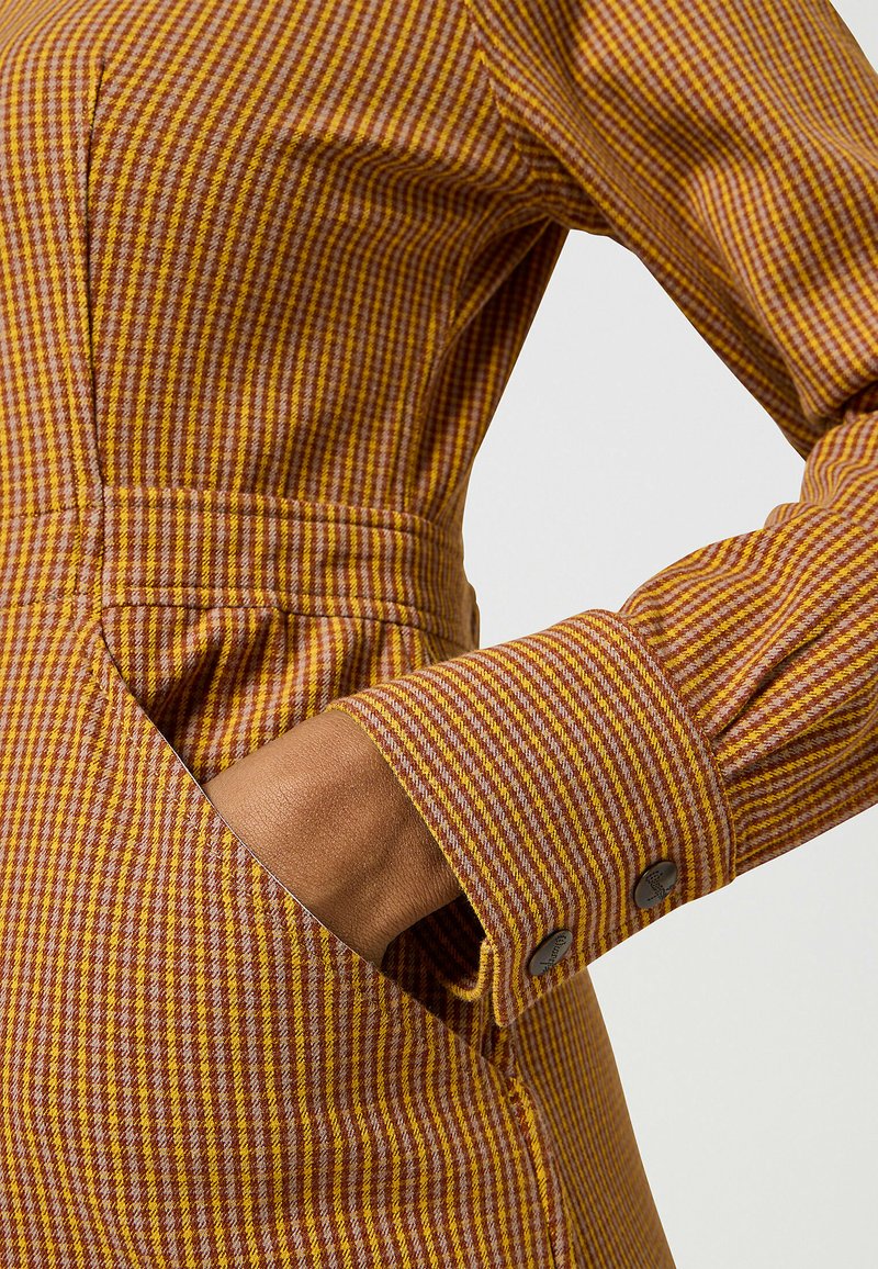 Abito a quadratini marroni e gialli con maniche lunghe, caratterizzato da un design aderente, tasche laterali e polsini con bottone. La texture sembra liscia.