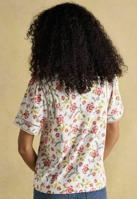 Krótki rękaw biały top z kwiatowym wzorem w odcieniach różu i niebieskiego, z koronkowymi detalami przy rękawach i dekolcie. Swobodny krój.