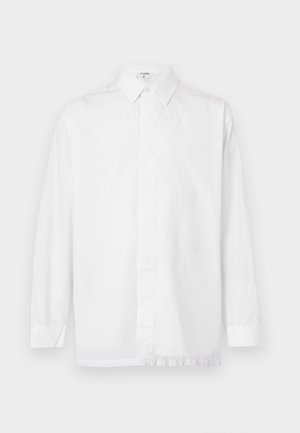 Jeune homme debout, les mains dans les poches, portant une chemise blanche à manches longues, un pantalon noir et des chaussures habillées noires sur un fond uni.