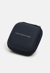 Kvadro zlatni neseser s glatkom vanjskom površinom. Na prednjoj strani središnje je smješten bijeli logo "TOMMY HILFIGER" u modernom fontu.