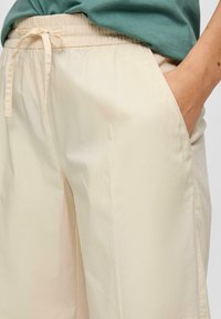 Pantalons légers de couleur crème, à coupe décontractée avec taille élastique et cordon de serrage, dotés de poches latérales et d'une texture lisse.