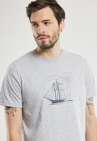 T-shirt en coton gris avec un graphique bleu marine d'un voilier, présentant des lignes géométriques et des accents circulaires, le tout sur un fond uni.