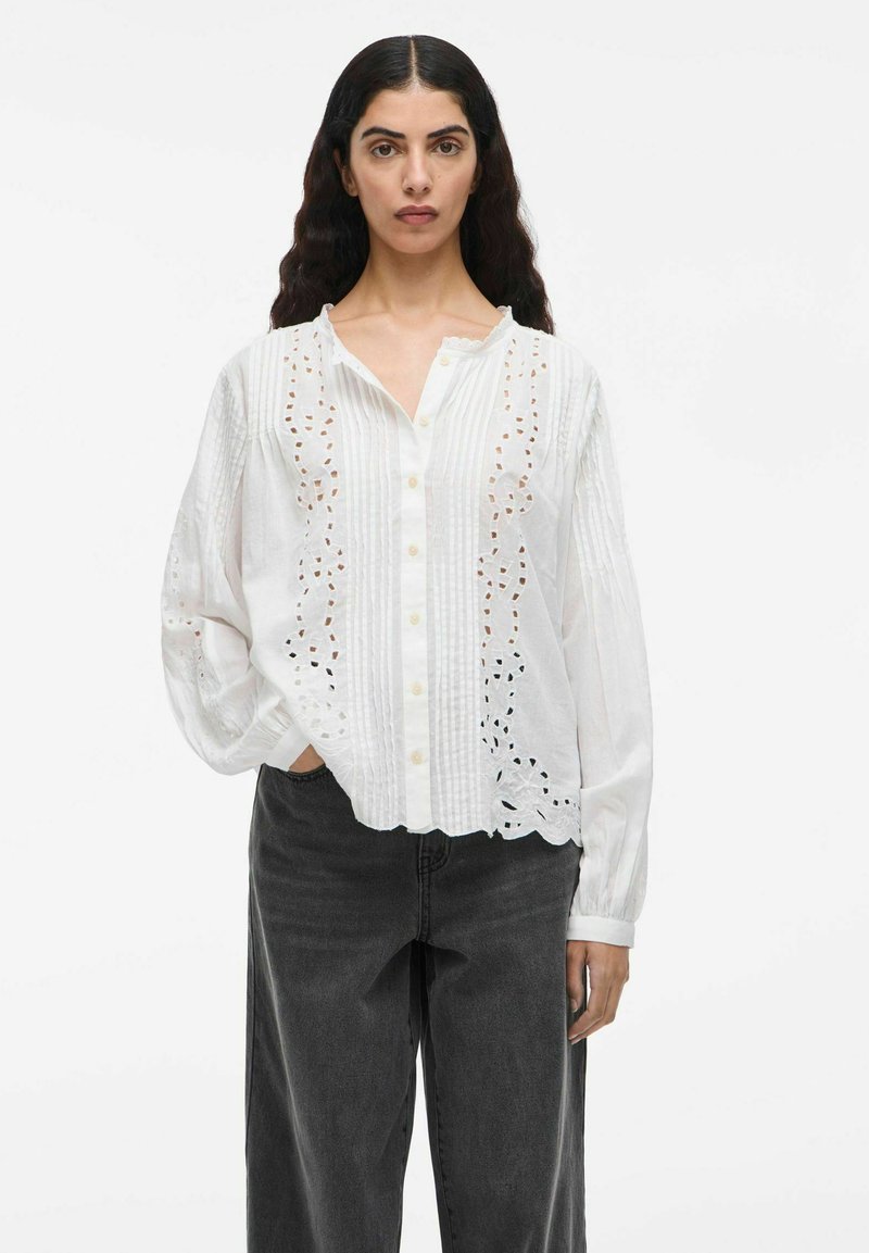 Weißes Hemd mit aufwendigen Ausschnitt-Designs, strukturierten Einsätzen und langen Ärmeln. Kombiniert mit schwarzen weiten Jeans.