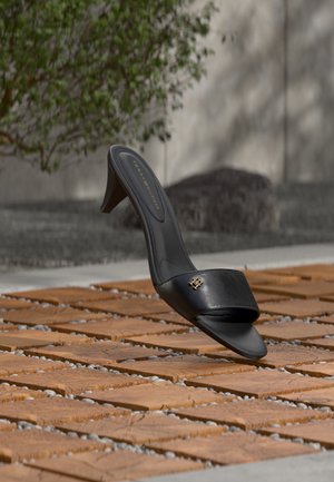 Μαύρο δερμάτινο πέδιλο με ανοιχτό δάχτυλο και τακούνι, με χρυσό λογότυπο στο λουράκι, που αιωρείται πάνω από ένα πλακιδωτό εξωτερικό χώρο με φυτά στο παρασκήνιο.