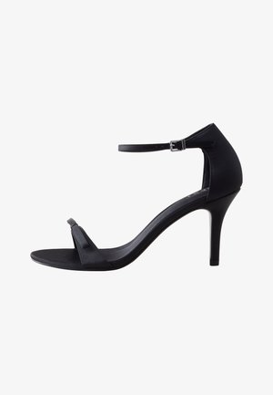 Sandale noire à talon haut avec fine bride à la cheville, bout ouvert et talon aiguille mince sur fond blanc.
