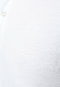 Polo bianco realizzato in tessuto testurizzato, caratterizzato da un unico bottone e un logo in rilievo sul petto.