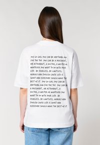 Witte katoenen t-shirt met grote zwarte bedrukte motiverende tekst op de achterkant. Korte mouwen en een losse pasvorm, met blauwe spijkerbroek zichtbaar.