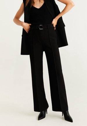 Frau trägt schwarze, hoch taillierte, weit geschnittene Hose mit Gürtel, schwarzen ärmellosen Top, Blazer über die Schultern gelegt und schwarze spitze High Heels.