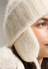Krémovo sfarbená vlnená čiapka s klapkami na uši, s jemným, textúrovaným povrchom a rebrovaným dizajnom. Materiál pôsobí teplo a útulne.