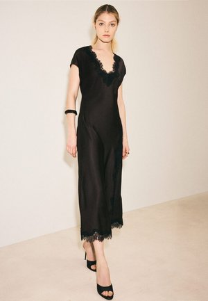 Frau in schwarzem ärmellosem Kleid mit Spitzenbesatz, trägt schwarze Sandalen mit hohen Absätzen und steht an einer beigen Wand und einem beigen Boden.