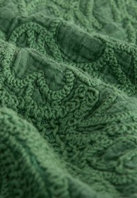 Tissu vert avec broderie en relief, texturée et bouclée, créant des motifs ondulés et géométriques sur une surface douce et froissée.