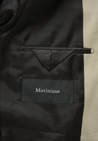 Tissu noir à texture lisse et finition brillante, avec une poche élégante et une étiquette portant le texte "Matinique" en broderie blanche.